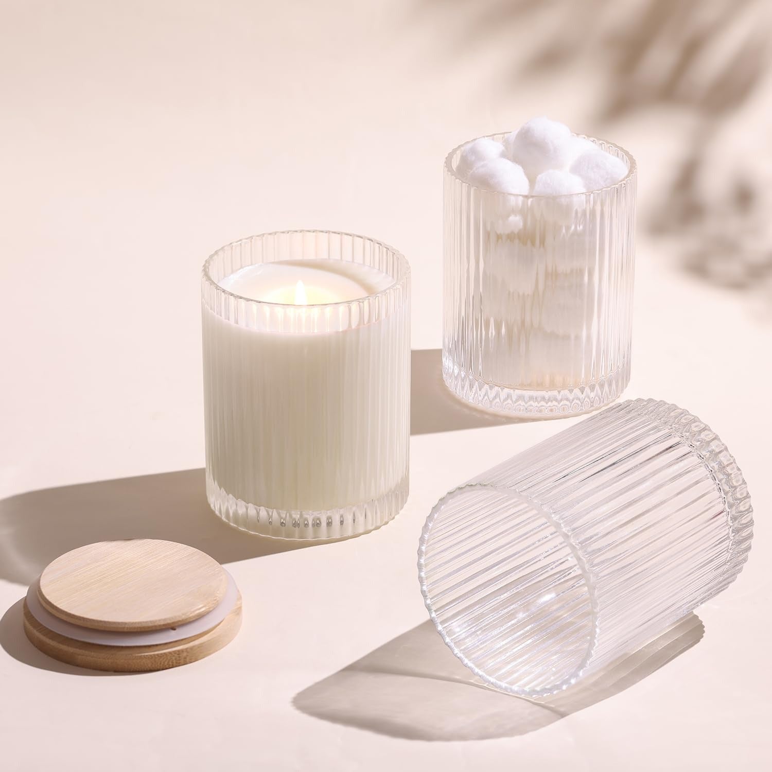 Three ribbed glass candle jars on beige surface, one with lit candle, one with cotton balls, bamboo lids beside