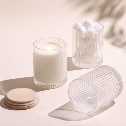 Three ribbed glass candle jars on beige surface, one with lit candle, one with cotton balls, bamboo lids beside