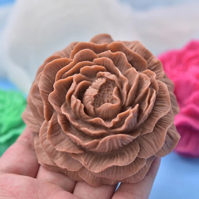 Hand holding large detailed peony flower candle cast in terracotta brown with layered petals, white and pink silicone moulds in background
