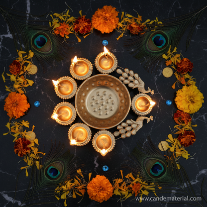 Overhead flat lay of gold peacock diya urli with lit diyas on dark marble with peacock feathers and gems