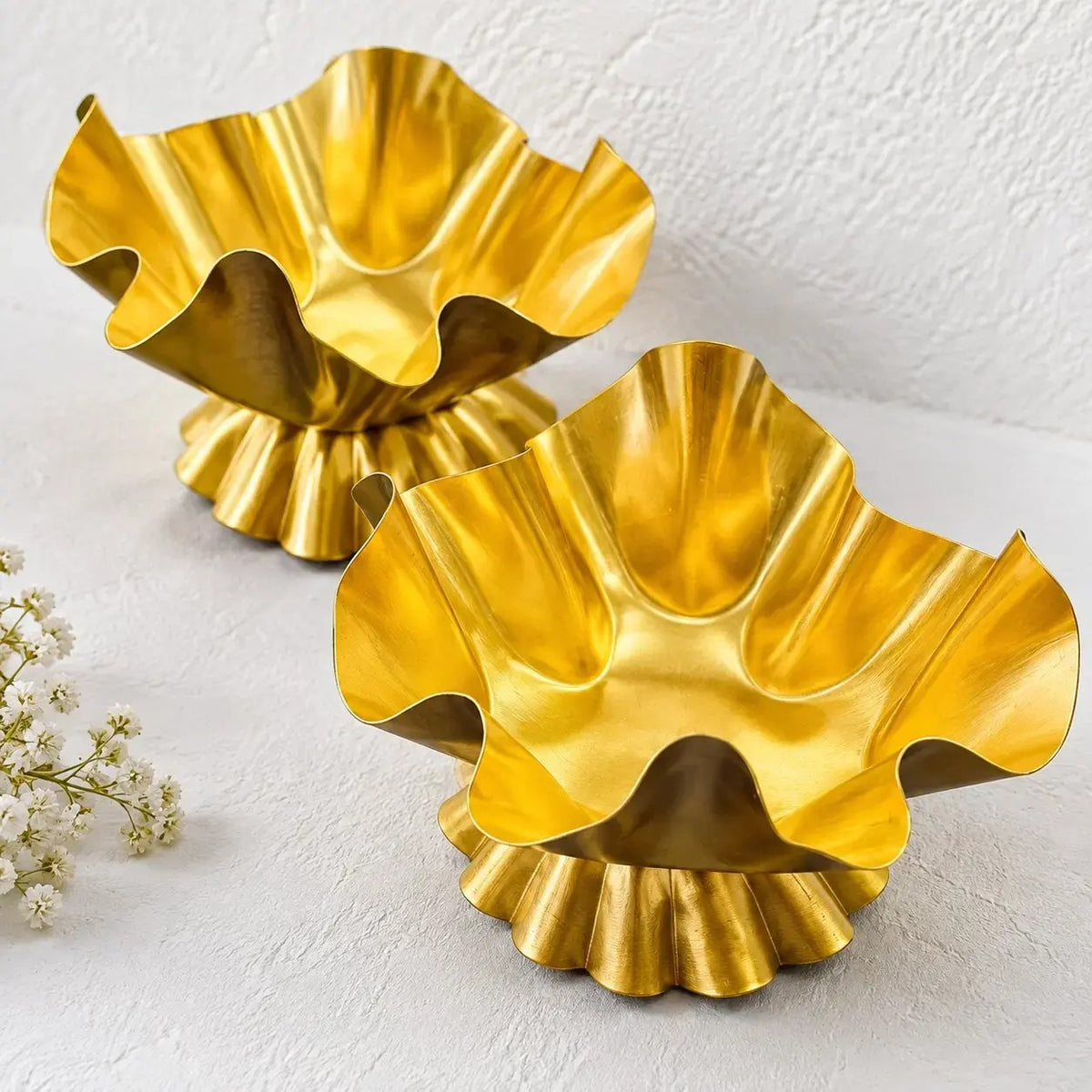 Two gold ruffled lotus petal freeform metal candle vessels on white textured surface with white baby's breath flowers